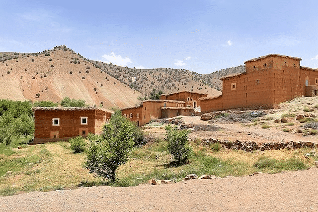 Des maisons traditionnelles en pisé rouge nichées au pied des montagnes arides du Haut Atlas sous un ciel bleu clair.