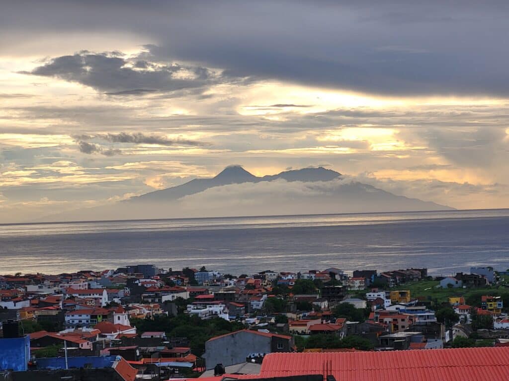 Vue panoramique de Tarrafal à Santiago au coucher du soleil, montrant les toits colorés et une église au premier plan, avec une imposante chaîne de montagnes émergeant des nuages à l'horizon au-dessus de l'océan Atlantique.