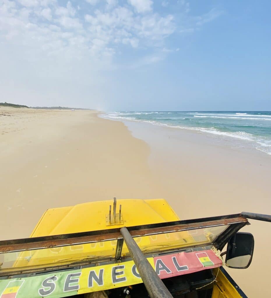 Vue depuis un véhicule 4x4 jaune parcourant une immense plage de sable fin bordée par l'océan au Sénégal.