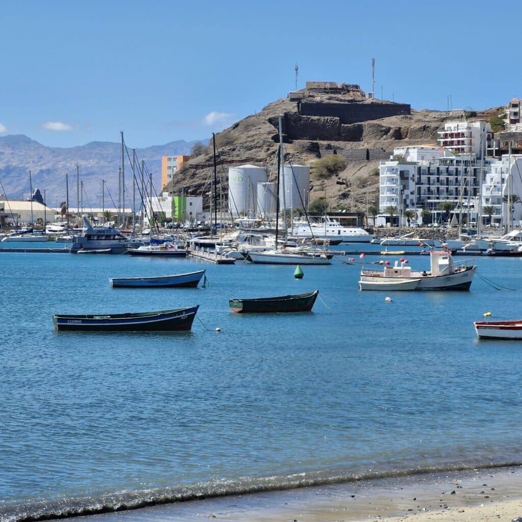 Vue du port de Mindelo à Sao Vicente avec des barques colorées, des voiliers et une colline fortifiée en arrière-plan.