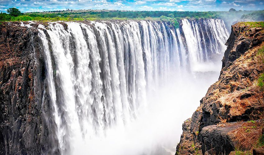 Vue panoramique des chutes Victoria avec d'immenses rideaux d'eau tombant dans un gouffre brumeux.