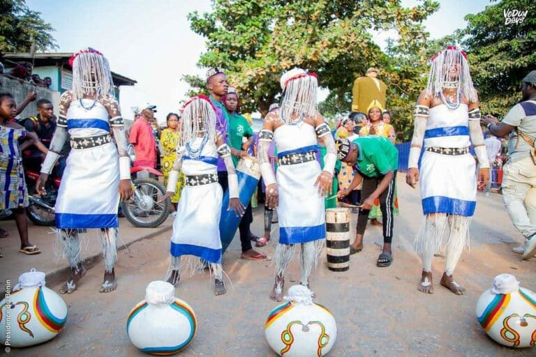 Quatre femmes en tenue blanche traditionnelle dansent lors d'une cérémonie de la fête du Vodoun au Bénin. Elles sont ornées de colliers de perles et exécutent des pas rituels sur un sol en terre battue.