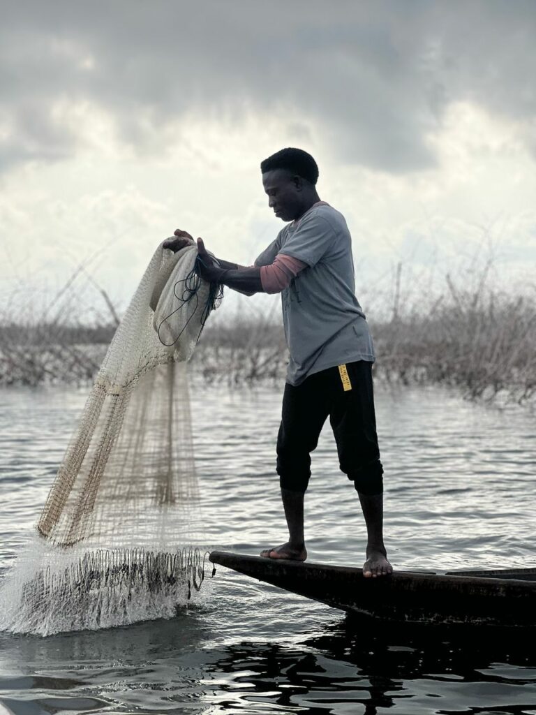 Pêcheur à Ganvié, au Bénin, lançant un filet depuis une pirogue traditionnelle sur le lac Nokoué, sous un ciel nuageux.