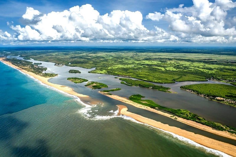 Vue aérienne spectaculaire d'une lagune côtière avec une langue de sable dorée séparant l'océan Atlantique et une rivière, sous un ciel partiellement nuageux, au Bénin.