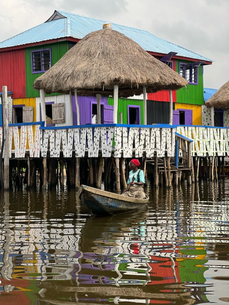 Femme en pirogue sur le lac Nokoué à Ganvié, au Bénin, devant des maisons colorées sur pilotis et une case traditionnelle au toit de chaume.