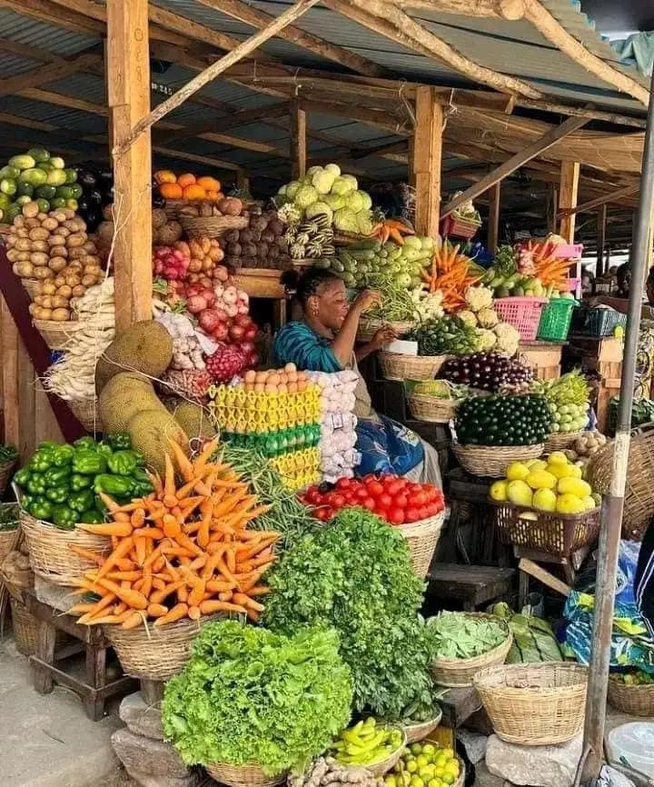 Une vendeuse assise derrière un étal coloré rempli de légumes et de fruits frais soigneusement disposés dans des paniers.