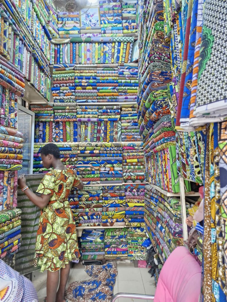 Une femme dans une boutique de pagnes colorés, entourée de centaines de rouleaux de tissu aux motifs africains.