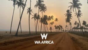 Route de sable bordée de palmiers au bord de l’océan Atlantique, avec un coucher de soleil à l’horizon. Logo blanc "WA AFRICA" au centre de l’image.