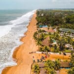 Vue aérienne de la plage de Grand-Bassam en Côte d’Ivoire, avec son sable doré, ses vagues blanches qui viennent s’écraser sur le rivage et ses rangées de cocotiers bordant des bâtiments et restaurants en bord de mer.