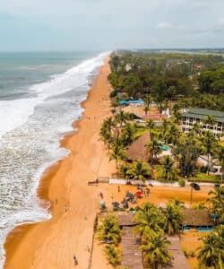 Vue aérienne de la plage de Grand-Bassam en Côte d’Ivoire, avec son sable doré, ses vagues blanches qui viennent s’écraser sur le rivage et ses rangées de cocotiers bordant des bâtiments et restaurants en bord de mer.