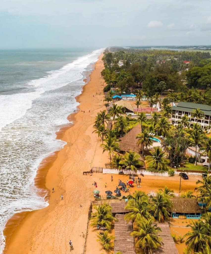 Vue aérienne de la plage de Grand-Bassam en Côte d’Ivoire, avec son sable doré, ses vagues blanches qui viennent s’écraser sur le rivage et ses rangées de cocotiers bordant des bâtiments et restaurants en bord de mer.