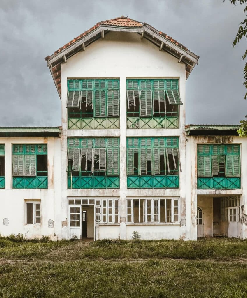 Bâtiment colonial abandonné à Grand-Bassam, Côte d’Ivoire, avec volets verts usés et peinture écaillée, sous un ciel gris orageux.