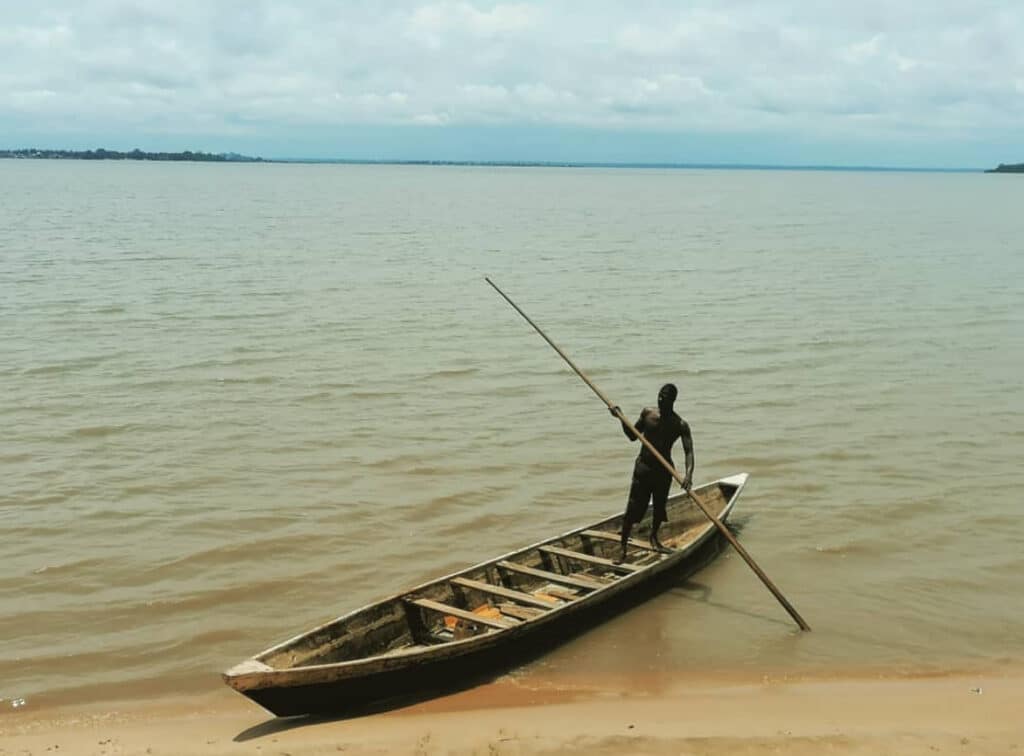 Homme debout dans une pirogue traditionnelle sur le lac Togo, tenant une longue perche pour naviguer.