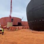 Chantier du futur musée du Vaudou à Porto-Novo, avec des bâtiments circulaires en construction, une structure noire imposante et une grue rouge sur un sol ocre.