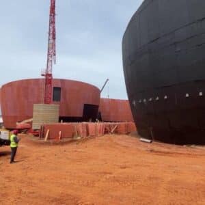 Chantier du futur musée du Vaudou à Porto-Novo, avec des bâtiments circulaires en construction, une structure noire imposante et une grue rouge sur un sol ocre.