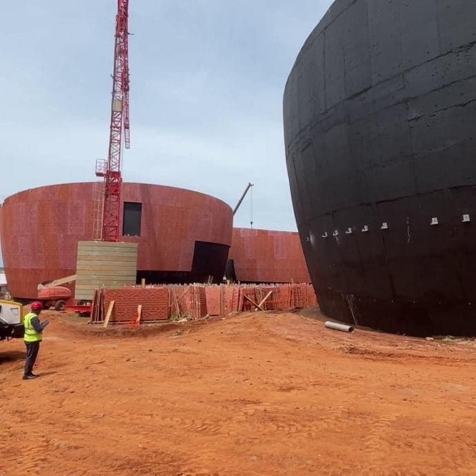 Chantier du futur musée du Vaudou à Porto-Novo, avec des bâtiments circulaires en construction, une structure noire imposante et une grue rouge sur un sol ocre.