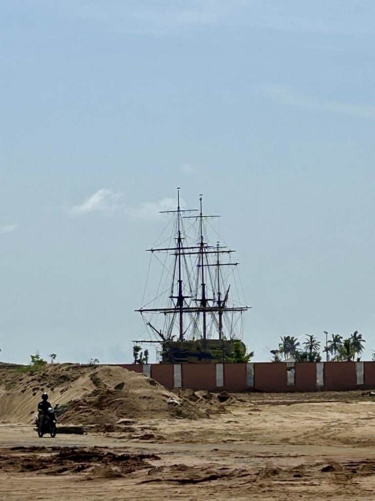 Réplique du navire historique à Ouidah au Bénin, vue depuis une zone sablonneuse avec un motocycliste au premier plan