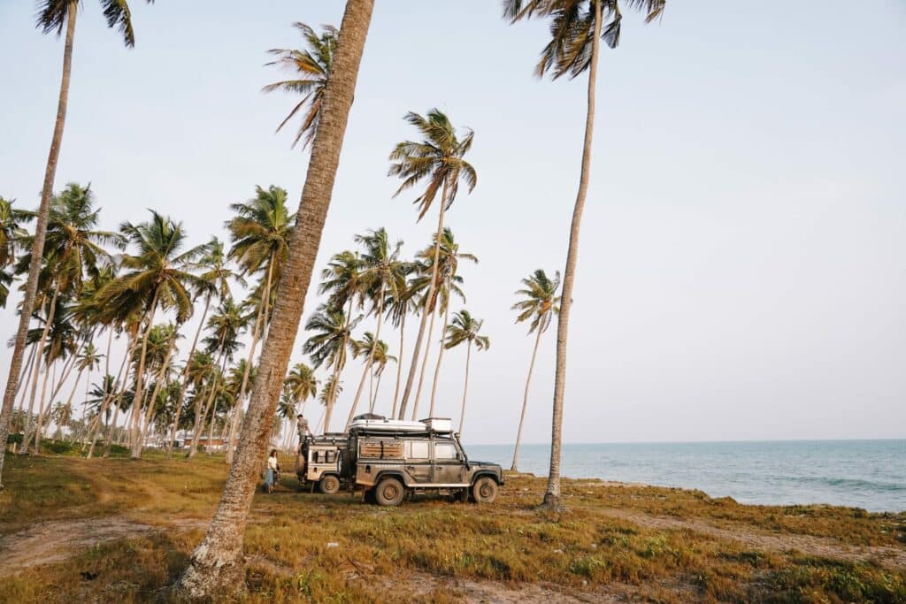 Véhicules 4x4 de type Land Rover Defender stationnés sous des palmiers au bord de l'océan lors d'un road trip entre le Bénin et le Ghana.
