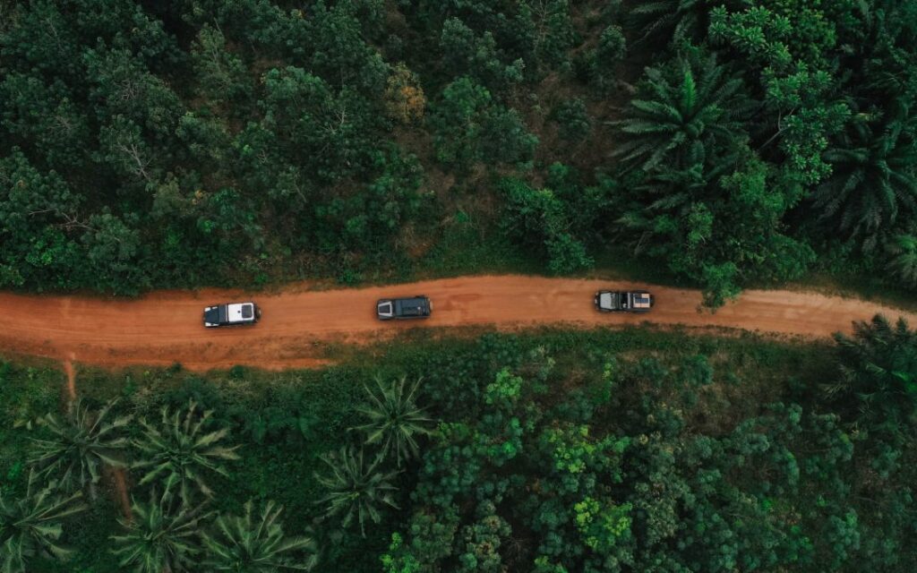 Vue aérienne par drone d'un convoi de véhicules 4x4 sur une route de terre rouge traversant une forêt dense lors d'un road trip entre le Bénin et le Ghana.