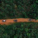 Vue aérienne par drone d'un convoi de véhicules 4x4 sur une route de terre rouge traversant une forêt dense lors d'un road trip entre le Bénin et le Ghana.