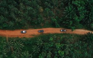 Vue aérienne par drone d'un convoi de véhicules 4x4 sur une route de terre rouge traversant une forêt dense lors d'un road trip entre le Bénin et le Ghana.