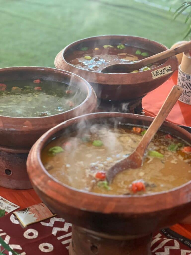 Trois pots en terre cuite contenant des sauces traditionnelles béninoises fumantes, dont une sauce tomate, servies lors d'un buffet.