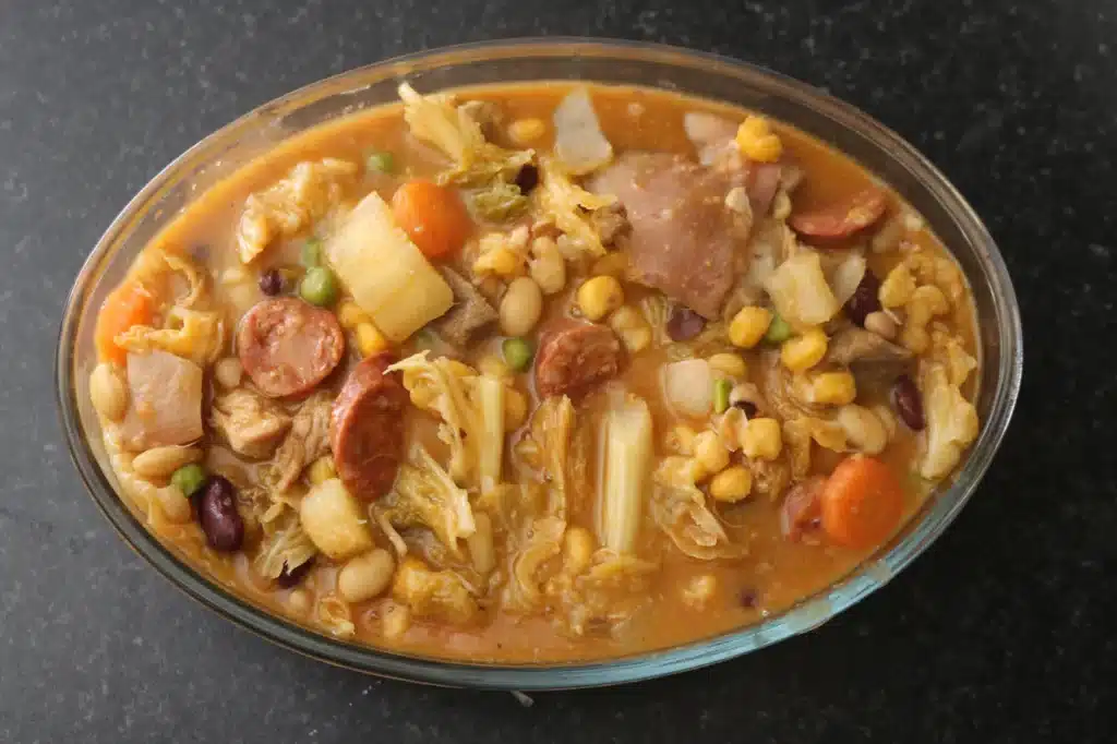 Un bol en verre rempli de Cachupa, ragoût traditionnel du Cap-Vert, composé de maïs, de haricots, de morceaux de viande, de tranches de saucisse (chorizo) et de légumes comme des carottes et du chou.