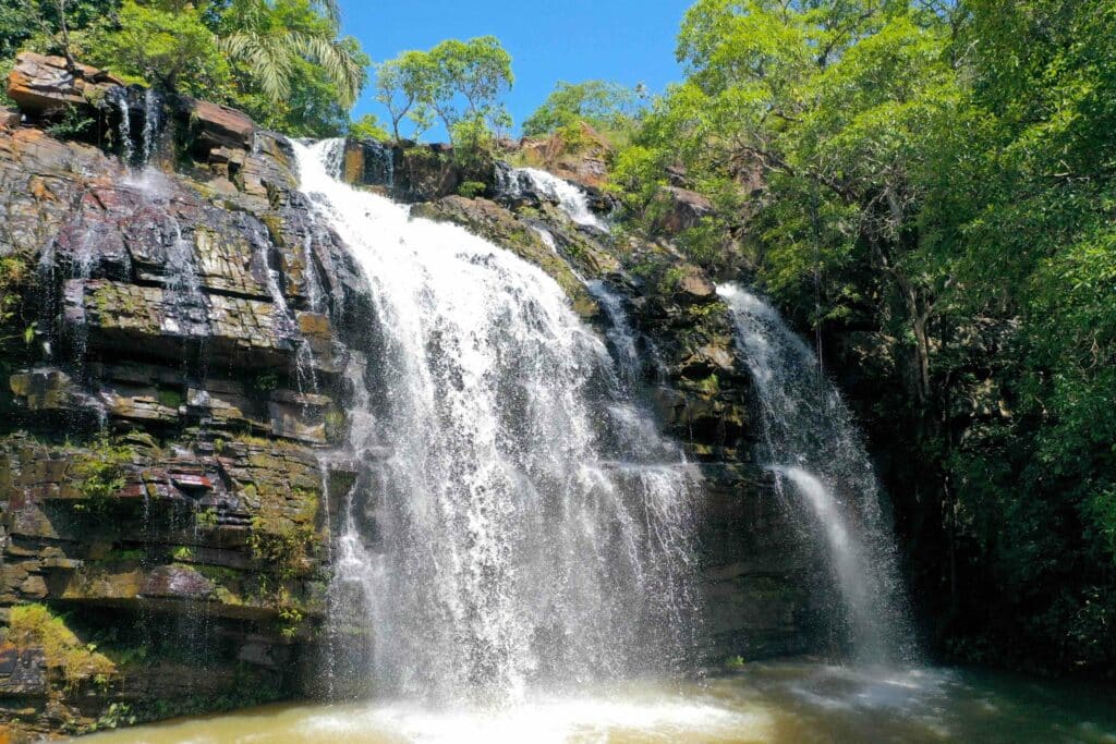 Impressionnante cascade d'eau s'écoulant sur plusieurs niveaux de parois rocheuses sombres, entourée d'une végétation tropicale luxuriante et d'arbres verts sous un ciel bleu clair, Bénin.