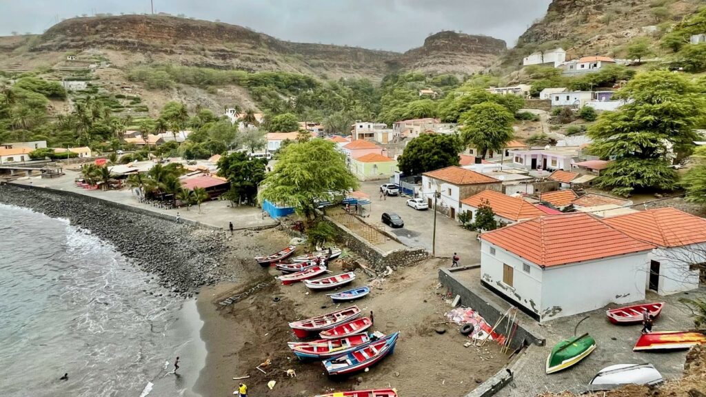 Vue du front de mer de Cidade Velha au Cap-Vert, montrant des barques de pêche colorées sur une plage de sable noir, des maisons aux toits de tuiles rouges et des falaises escarpées en arrière-plan par temps couvert.