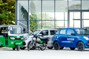 Alignement de différents véhicules de transport Gozem devant un bâtiment moderne vitré : un tricycle vert et blanc (tuk-tuk), une moto taxi et deux citadines (une grise et une bleue) arborant le logo de l'application.