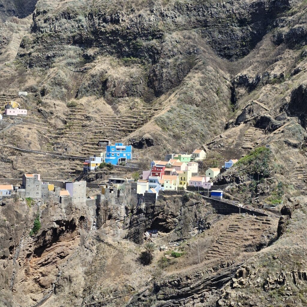 Vue aérienne spectaculaire d'un petit village de montagne aux maisons colorées (bleues, rouges) niché au pied de falaises volcaniques immenses et arides, avec des cultures en terrasses sculptées à flanc de colline dans la Vallée de Paul, Santo Antão.