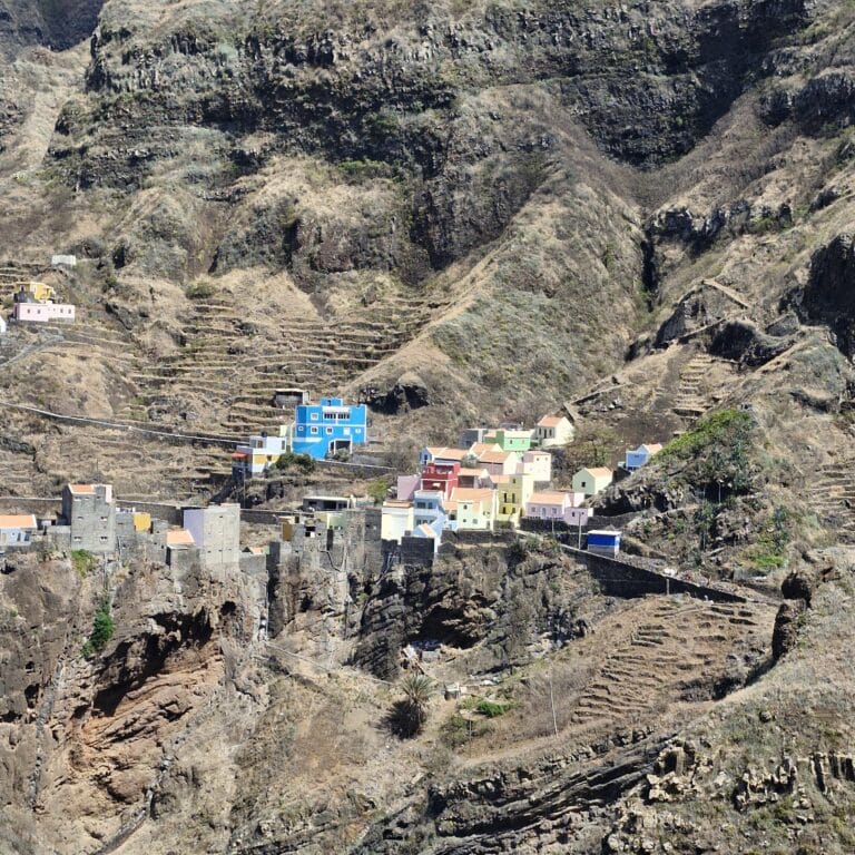 Vue aérienne spectaculaire d'un petit village de montagne aux maisons colorées (bleues, rouges) niché au pied de falaises volcaniques immenses et arides, avec des cultures en terrasses sculptées à flanc de colline dans la Vallée de Paul, Santo Antão.