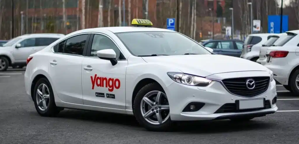 Voiture berline blanche de marque Mazda arborant le logo rouge Yango sur la portière avant, stationnée dans un parking extérieur avec un panneau "Taksi" sur le toit.