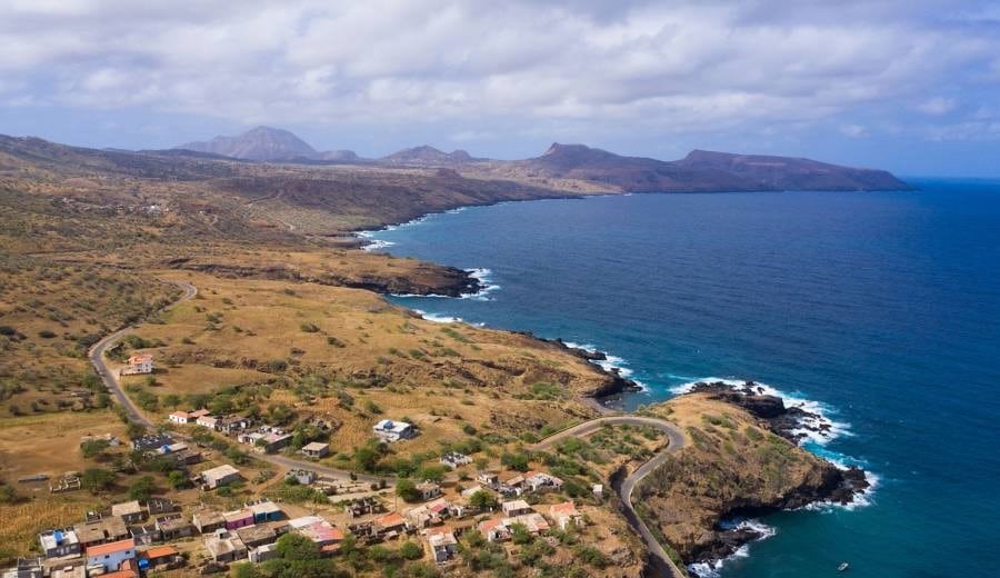 Vue aérienne de la côte escarpée de l'île de Santiago au Cap-Vert, montrant un petit village aux maisons colorées, une route côtière sinueuse et l'océan Atlantique bleu profond sous un ciel nuageux.