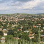 Vue panoramique de la ville de Natitingou au Bénin, montrant des habitations aux toits de tôle nichées dans une végétation dense au pied des collines de l'Atacora sous un ciel nuageux.