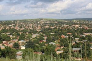 Vue panoramique de la ville de Natitingou au Bénin, montrant des habitations aux toits de tôle nichées dans une végétation dense au pied des collines de l'Atacora sous un ciel nuageux.