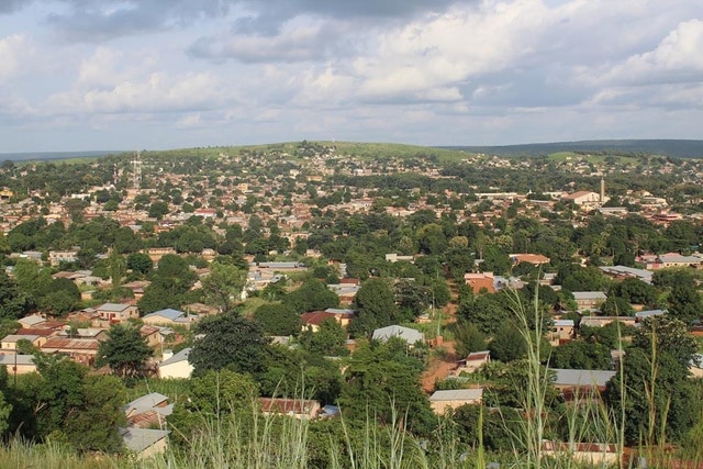 Vue panoramique de la ville de Natitingou au Bénin, montrant des habitations aux toits de tôle nichées dans une végétation dense au pied des collines de l'Atacora sous un ciel nuageux.