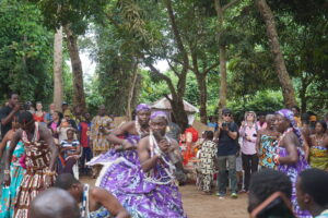 Une scène animée lors des Vodun Days à Ouidah, montrant des initiés en tenues violettes et bleues traditionnelles chantant et jouant des instruments devant un public de touristes et de locaux dans une forêt sacrée.