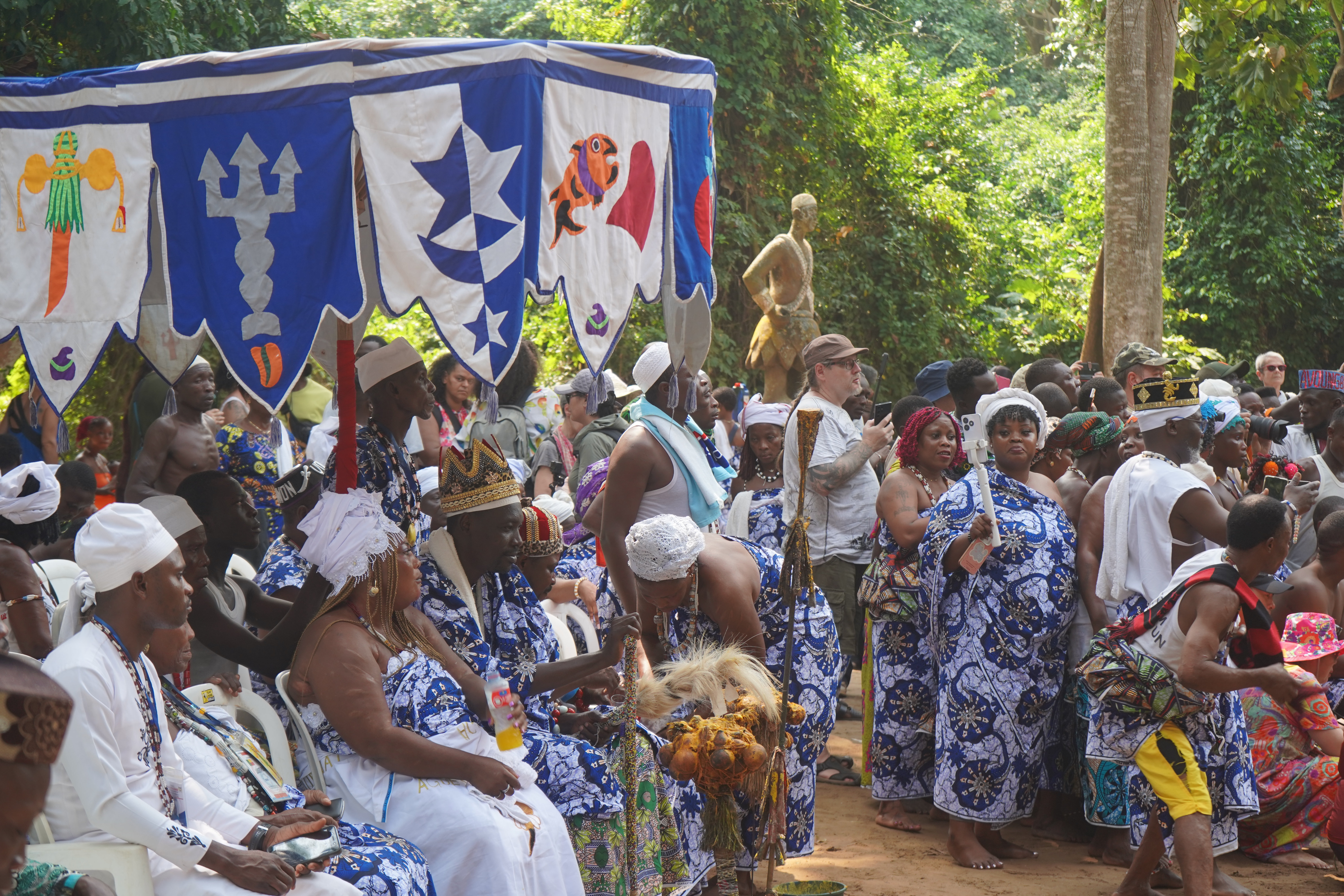 Une assemblée de dignitaires et de fidèles lors des Vodun Days à Ouidah. Des chefs traditionnels en tenues bleues et blanches brodées sont assis sous une grande ombrelle d'apparat ornée de symboles royaux (poisson, étoile, trident), entourés d'une foule dense dans un cadre boisé.