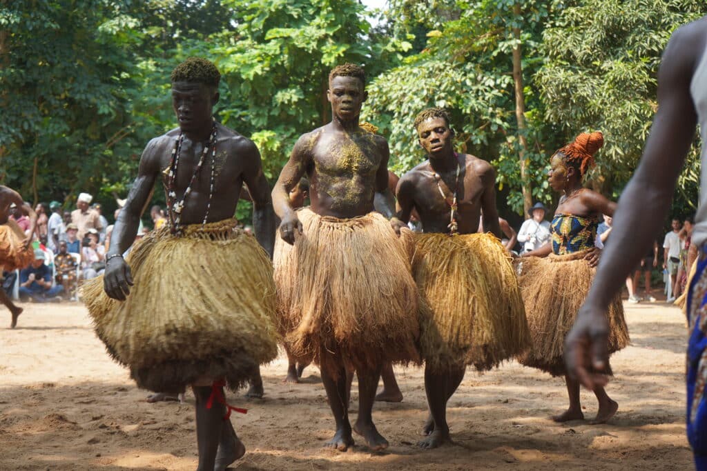 Quatre danseurs béninois, le buste nu et enduit de pigments, vêtus de jupes traditionnelles en fibres de raphia, avancent en rythme sur un sol de sable lors d'une cérémonie en plein air.