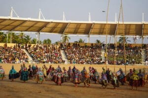 Une grande troupe de danseurs vêtus de costumes de masques Egungun colorés et richement brodés performe sur une place de sable devant des gradins remplis de spectateurs, sous une structure moderne, au Bénin.