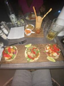Trois mini-tacos garnis de poisson et poivrons sur une planche en bois avec des olives et boissons en terrasse.