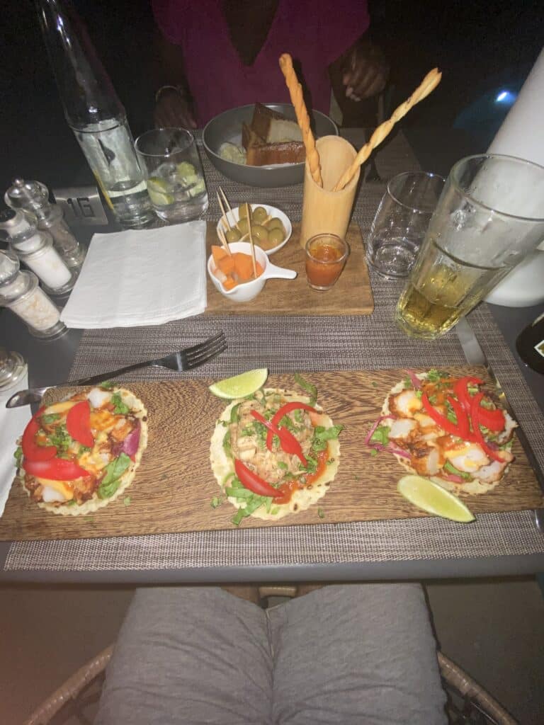 Trois mini-tacos garnis de poisson et poivrons sur une planche en bois avec des olives et boissons en terrasse.