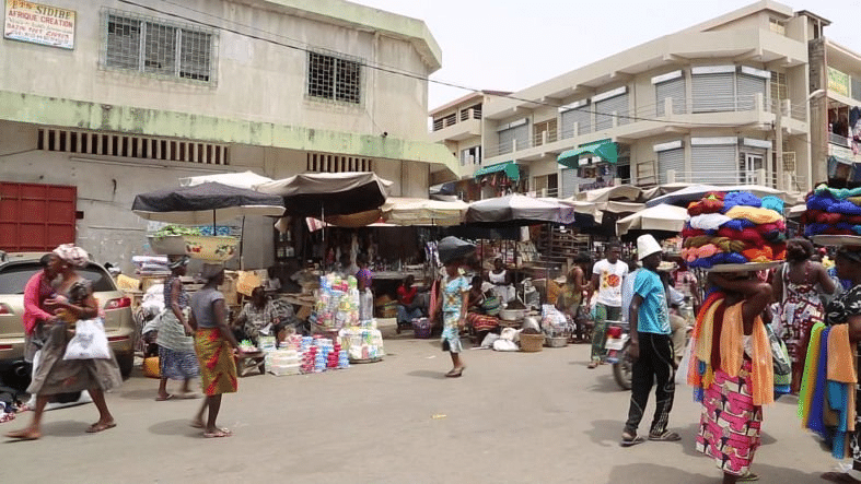 Scène de rue animée au Grand Marché d'Assigamé à Lomé, avec des commerçantes portant des marchandises sur la tête.