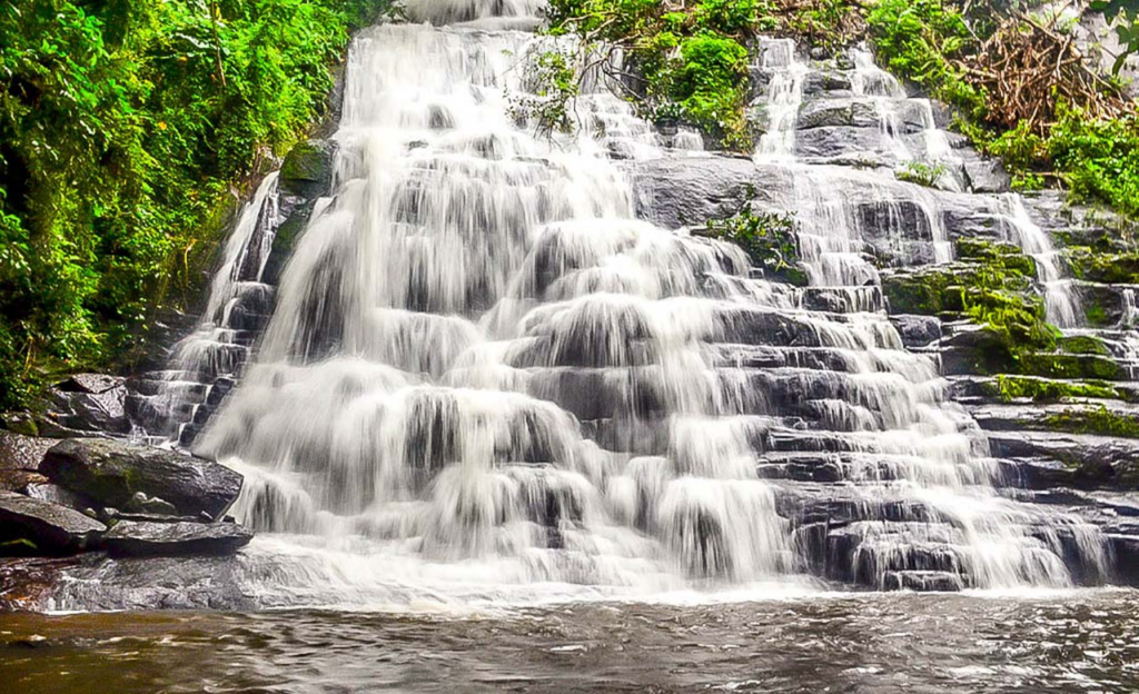 Une large cascade d'eau vive s'écoulant sur des rochers noirs en escalier entourés d'une forêt dense à Man.