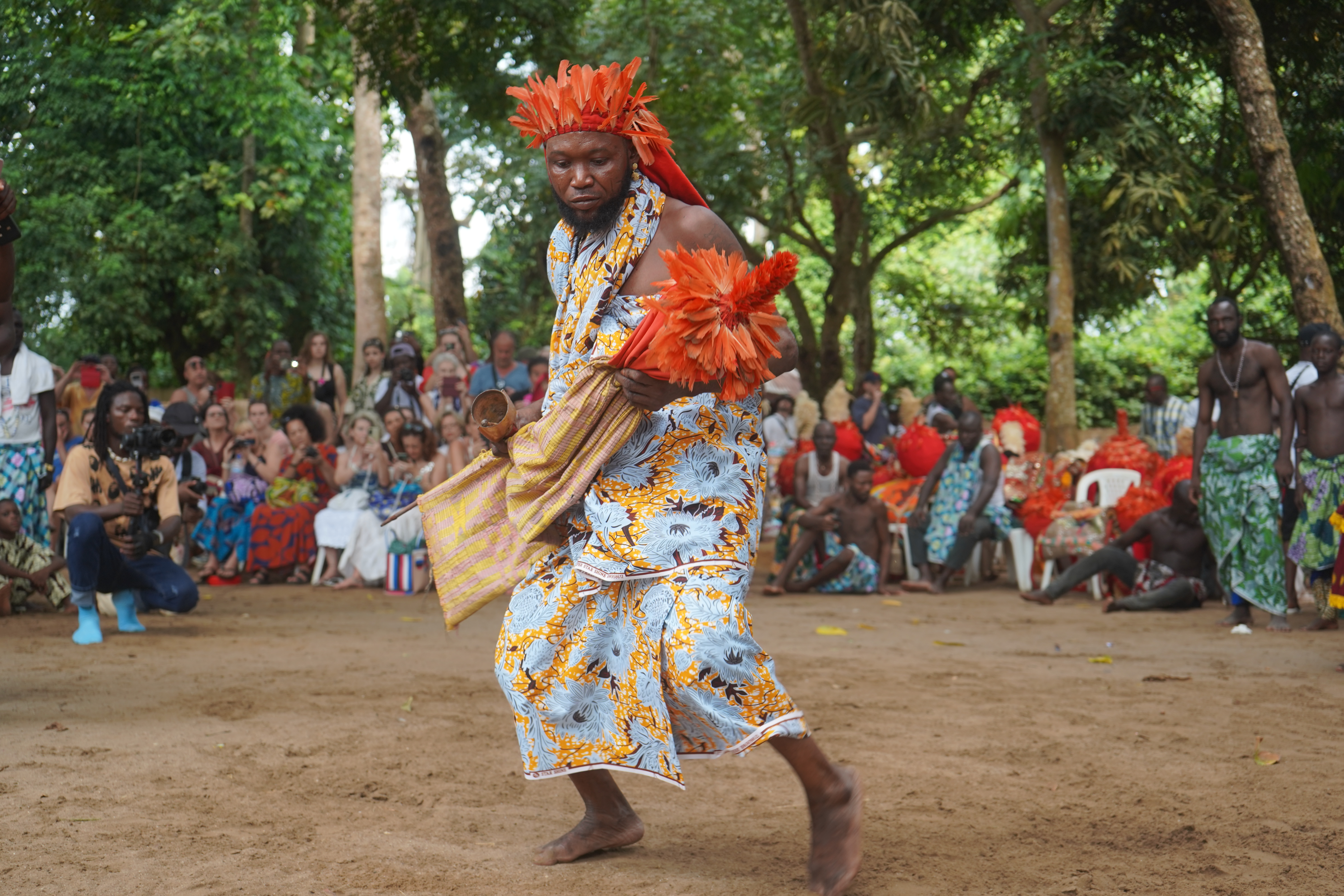 Un performeur béninois en pleine action lors des Vodun Days, portant une coiffe et des accessoires de plumes orange vif, vêtu d'un pagne traditionnel bleu et jaune à motifs, devant un public assis dans une zone boisée à Ouidah.
