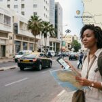 Une voyageuse afro-descendante avec sac à dos consulte une carte papier et son smartphone dans une rue moderne de Dakar, avec une incrustation de carte de l'Afrique de l'Ouest montrant le Sénégal, la Côte d'Ivoire, le Ghana et le Bénin avec des icônes de sécurité.