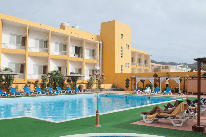 Piscine extérieure entourée de transats bleus devant la façade jaune de l'Hôtel Porto Grande au Cap-Vert.
