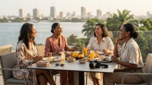 Quatre jeunes femmes afro-descendantes, élégantes et complices, partagent un déjeuner convivial sur une terrasse chic d'Abidjan, en Côte d'Ivoire. En arrière-plan, une vue panoramique sur la lagune Ébrié et le paysage urbain moderne du Plateau. Elles sont entourées de plats de gastronomie ivoirienne, de fruits tropicaux et d'un appareil photo.
